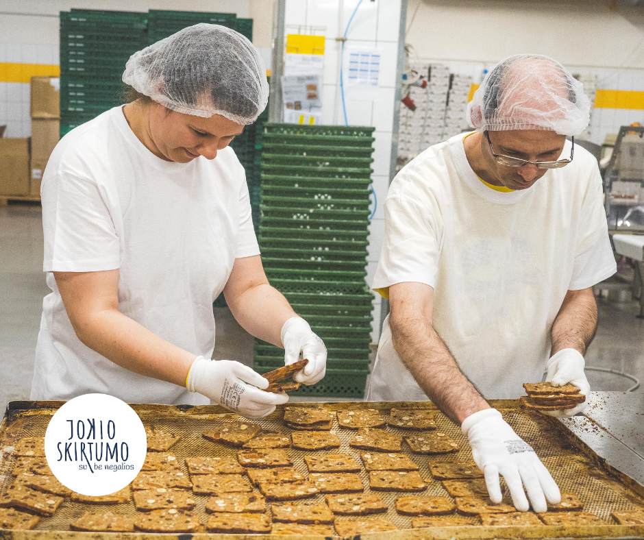 Nuotraukoje – du žmonės, dirbantys gamyboje, baltai apsirengę. Jie dėlioja duonos gaminius ant padėklo.